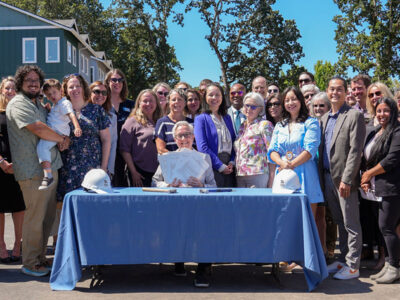 Governor Kotek sits behind a blue table in a Habitat neighborhood. She holds papers and is surrounded by smiling people.
