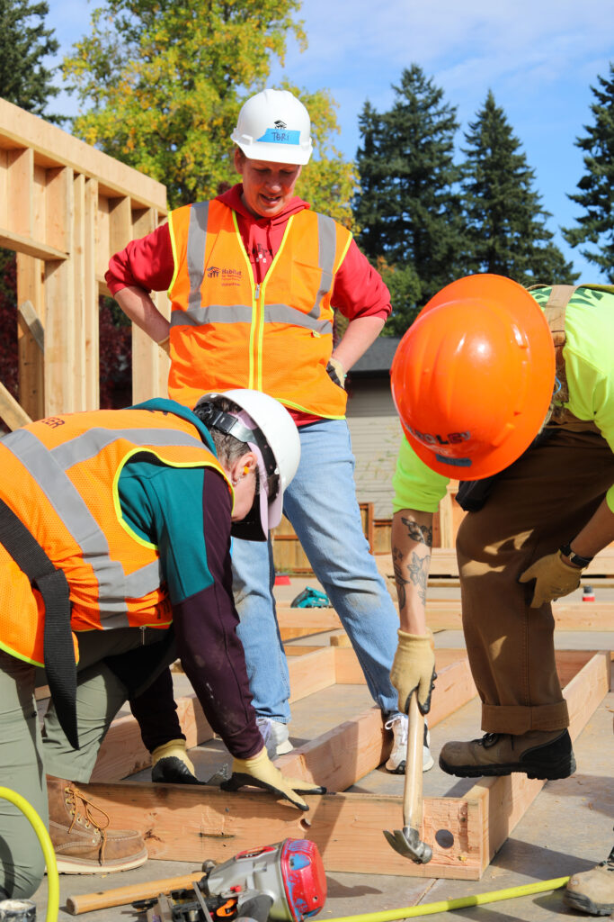 Three people in high-visibility vests and hardhats work together to frame a wall for a new Habitat home. The group of Habitat homebuyers and staff works together to hammer, secure beams, and guide the construction.