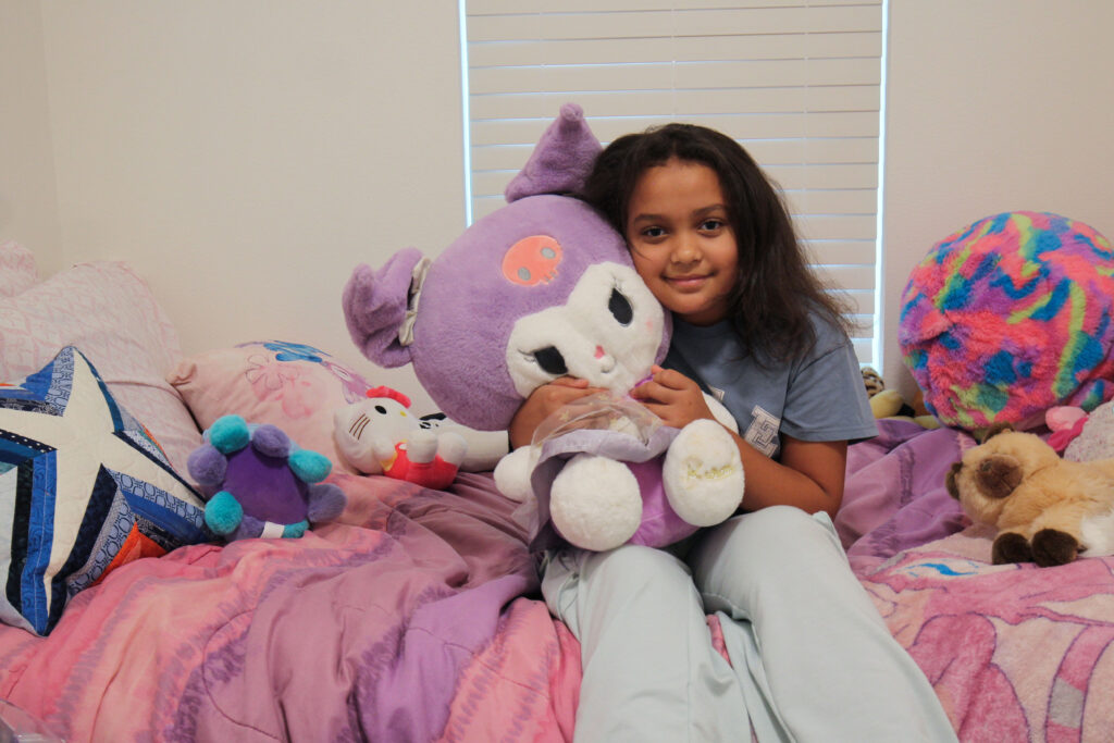 A child sits in her brightly decorated room in her Habitat home, hugging a stuffed animal.