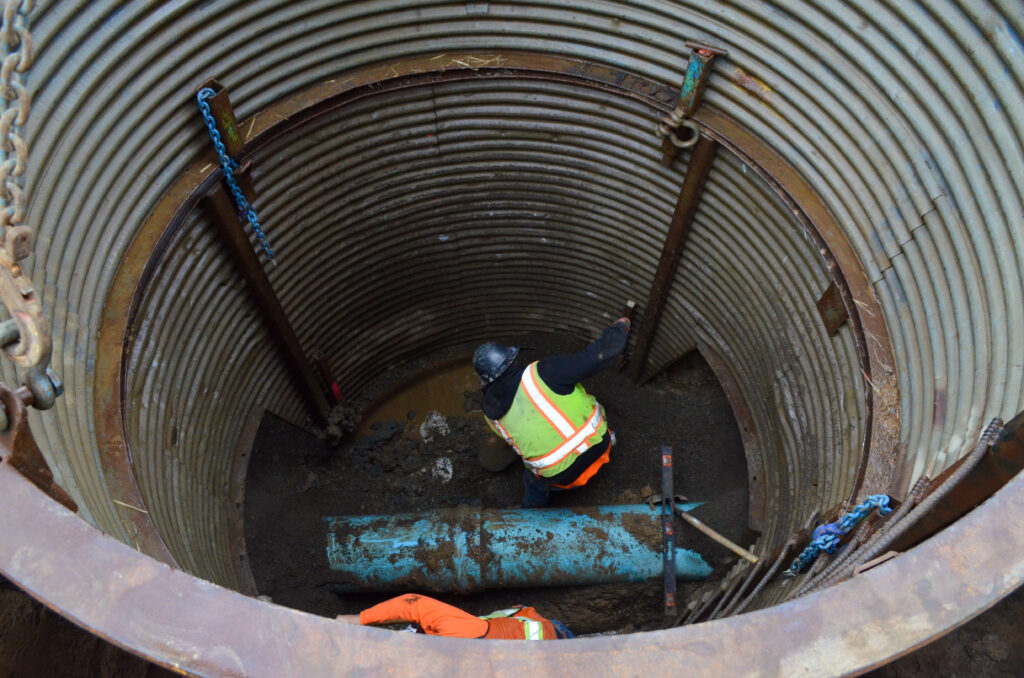 A Habitat crew member in a high-visibility vest and hardhat stands 15-feet below ground, digging to expose the water main and install a manhole for Gooseberry Trails.