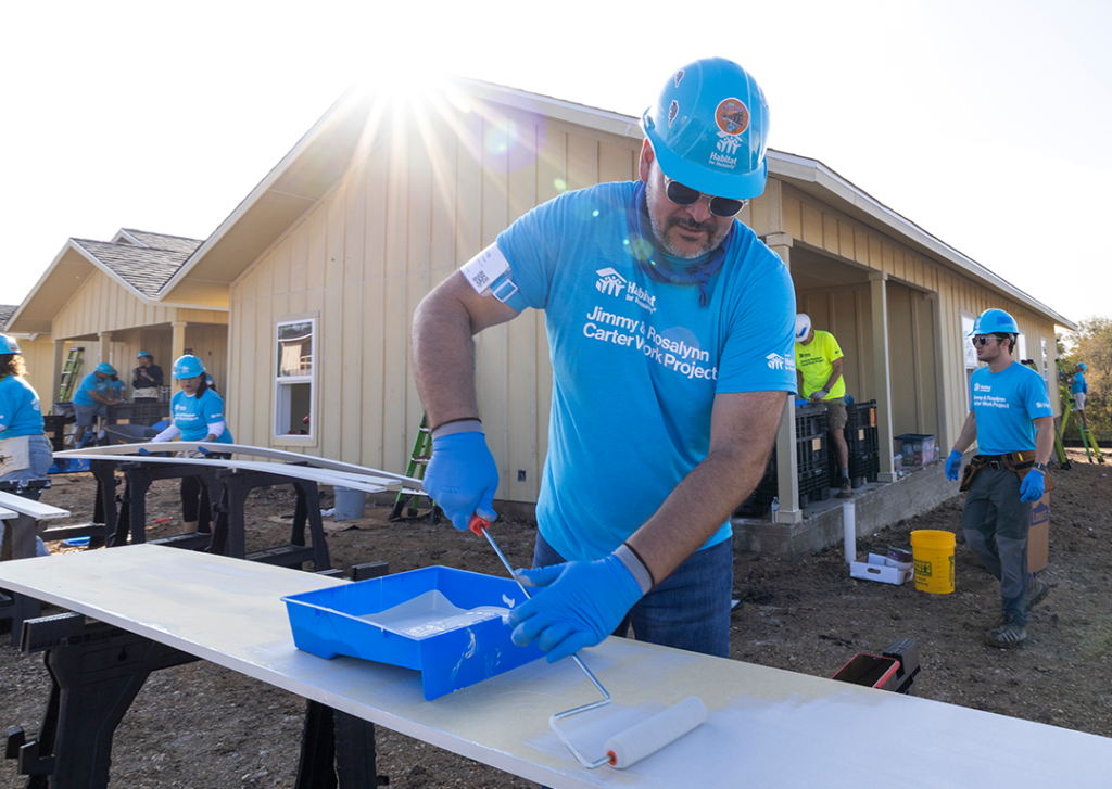 A Habitat volunteer wearing bright blue uses a roller to apply paint on a sunny job site. 