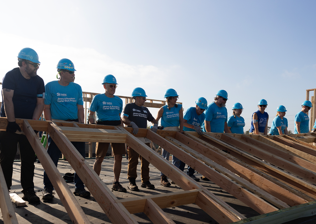 A dozen Habitat volunteers stand side by side to raise a framed wall on a clear day.  