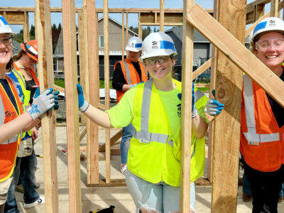 Sophia wear a high-visibility vest, hardhat, and safety goggles and smiles alongside two other ambassador council members, all holding up the framed wall of a Habitat house.