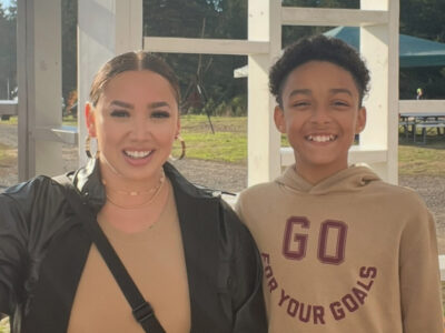 A Habitat homebuyer and her teenage son smile, looking content, standing outside on a sunny day.