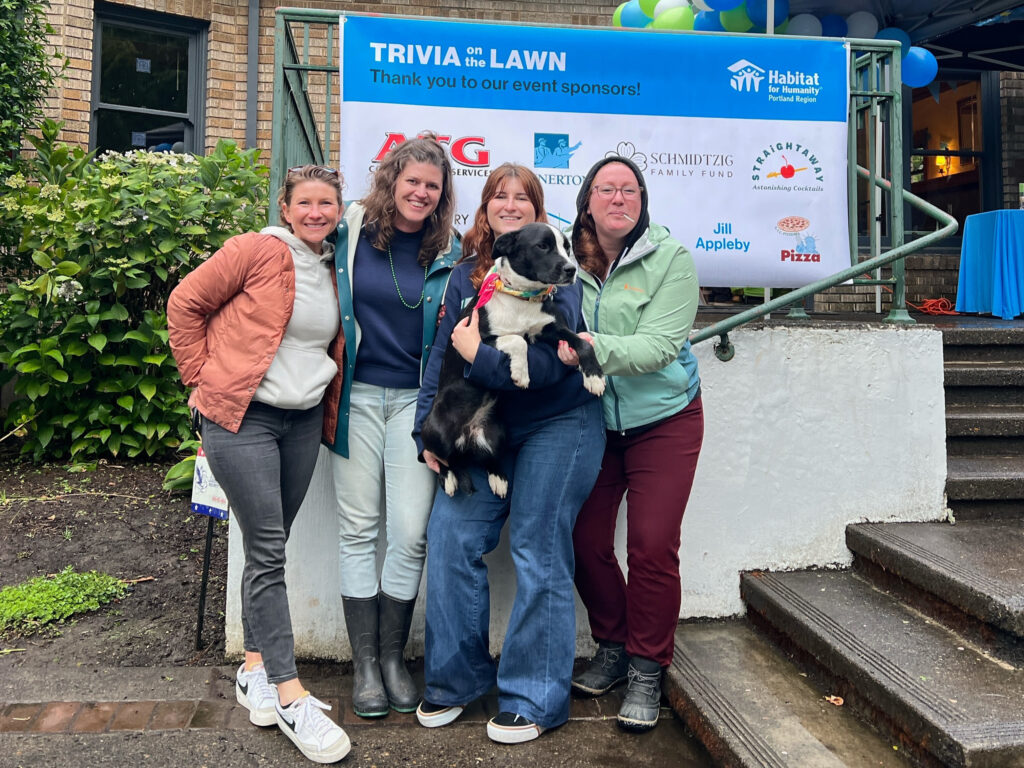 Sophia poses for a group photo in front of Habitat's "Trivia on the Lawn" sign with three smiling people, and she holds a black and white dog with a bandana in her arms. 
