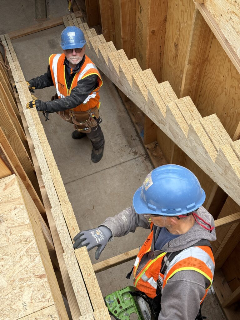 Ron works alongside another Core volunteer to frame a set of stairs at West Lake Grove. He looks up at the camera from inside the stairs-in-progress.