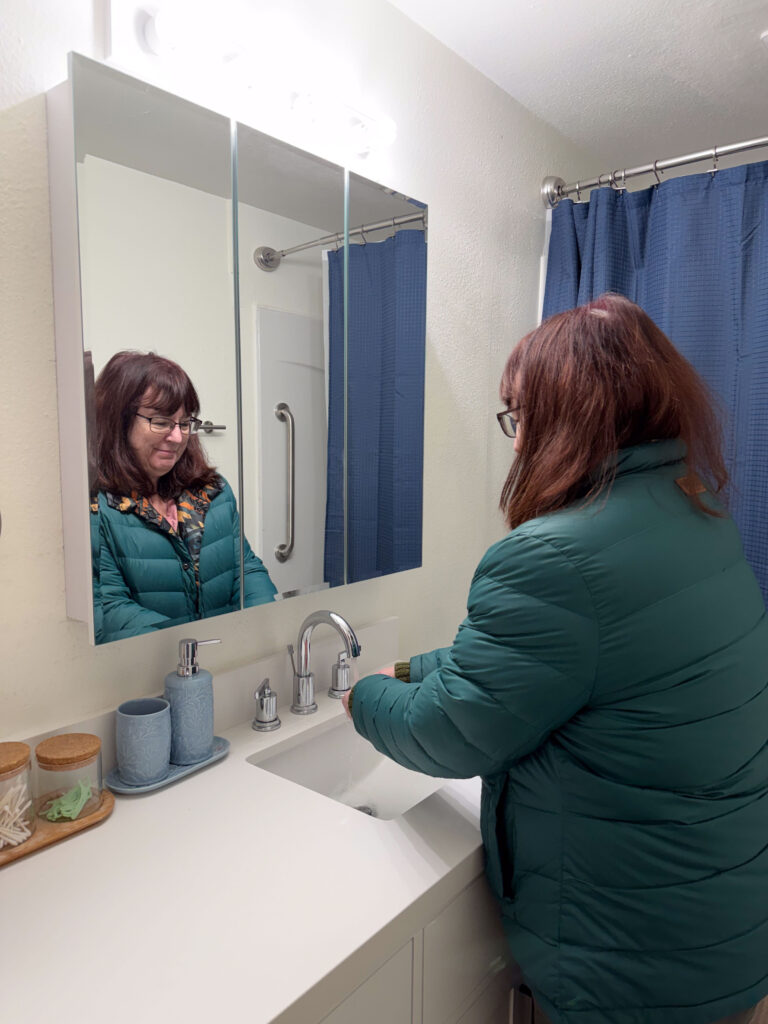 Habitat Home Repair client Sandra washes her hands in her brand new sink that was installed during her bathroom repair. She smiles in the mirror, surrounded by fresh white counters and walls.
