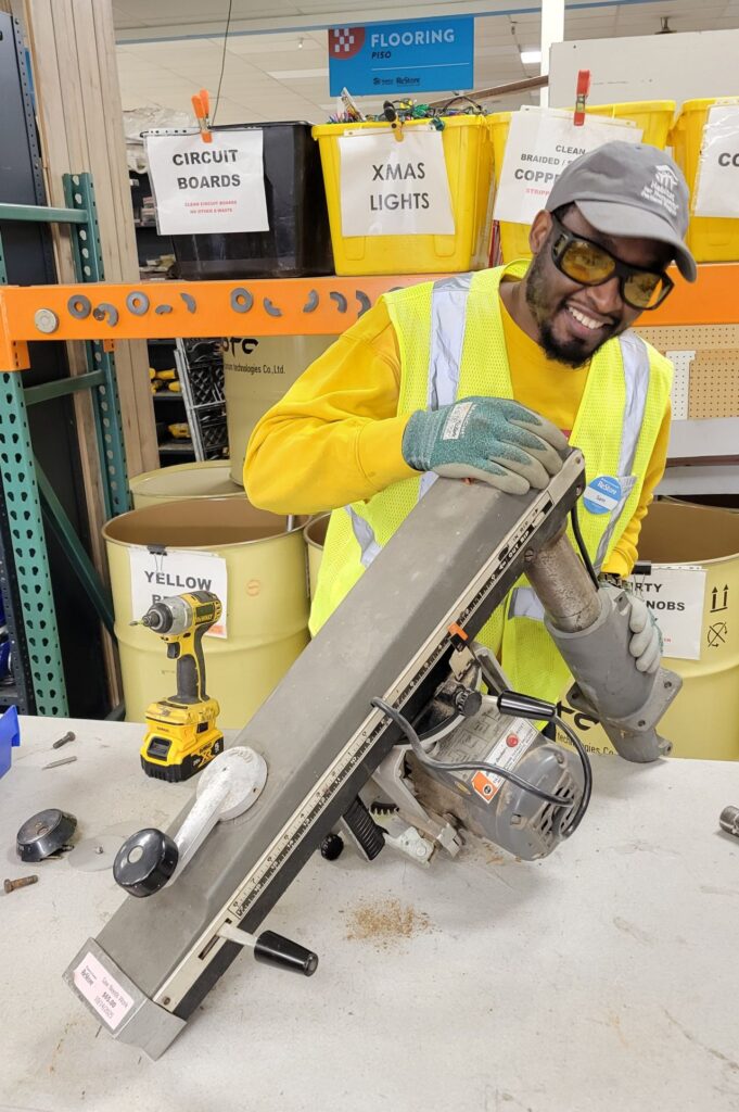 Samuel works on dismantling a table saw, wearing gloves, a reflective vest, and safety glasses.