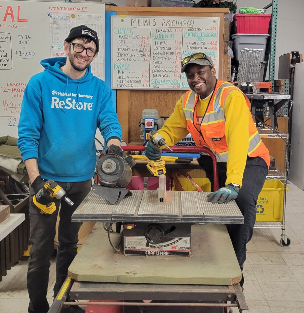 Samuel smiles alongside Habitat Salvage and Recycling Manager Joseph Drushal. Samuel holds a drill and shows off the remains of Craftsman saw from 1971 that he dismantled.