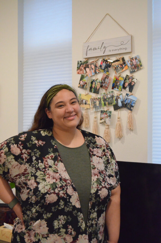 Stephanie smiles, standing in front of a sign that reads "family is everything" and boasts a collection of family photos.