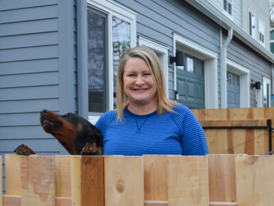 Habitat homebuyer Duffy grins in front of her new home, her dog beside her.