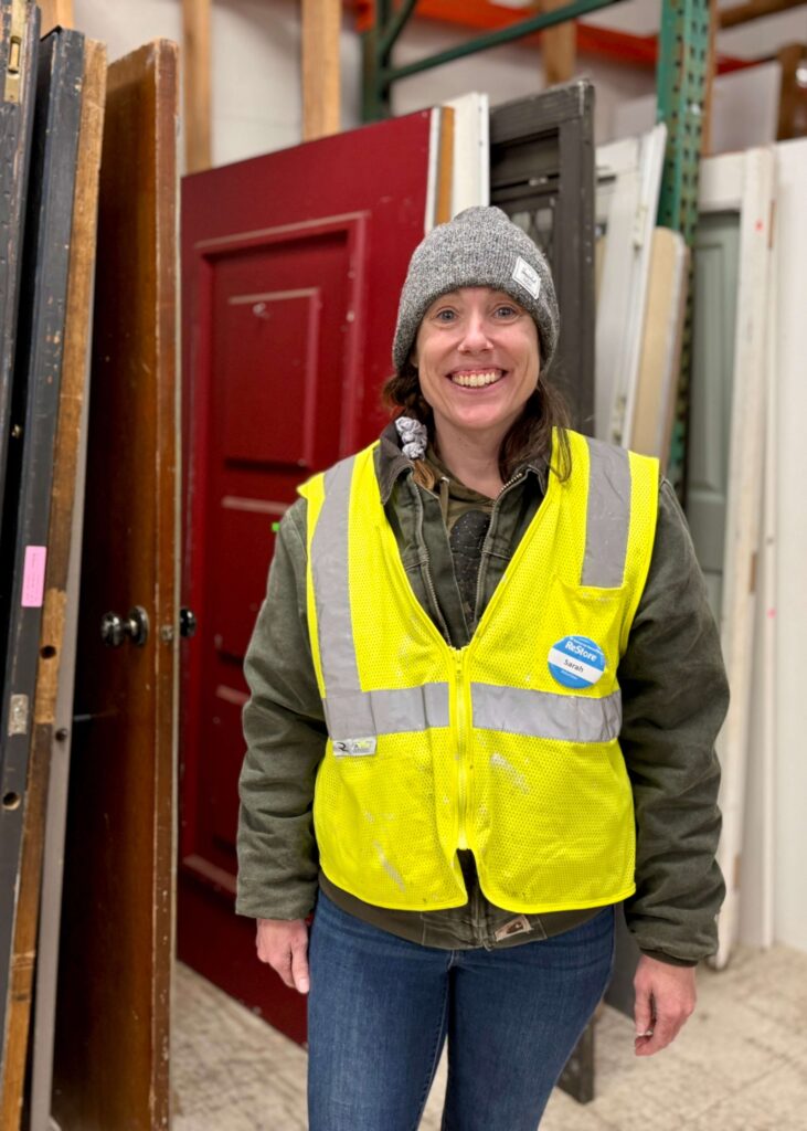 Sarah smiles in her Habitat volunteer vest and name badge, standing in front of a row of doors at the Gresham ReStore.